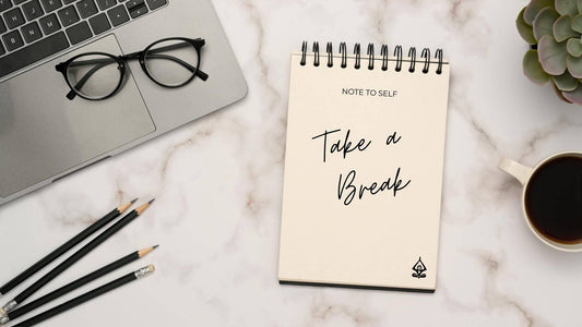 Flat lay of a calm workspace with a spiral notepad reading “note to self: take a break,” representing reducing stress at work and healthy stress management for professionals.